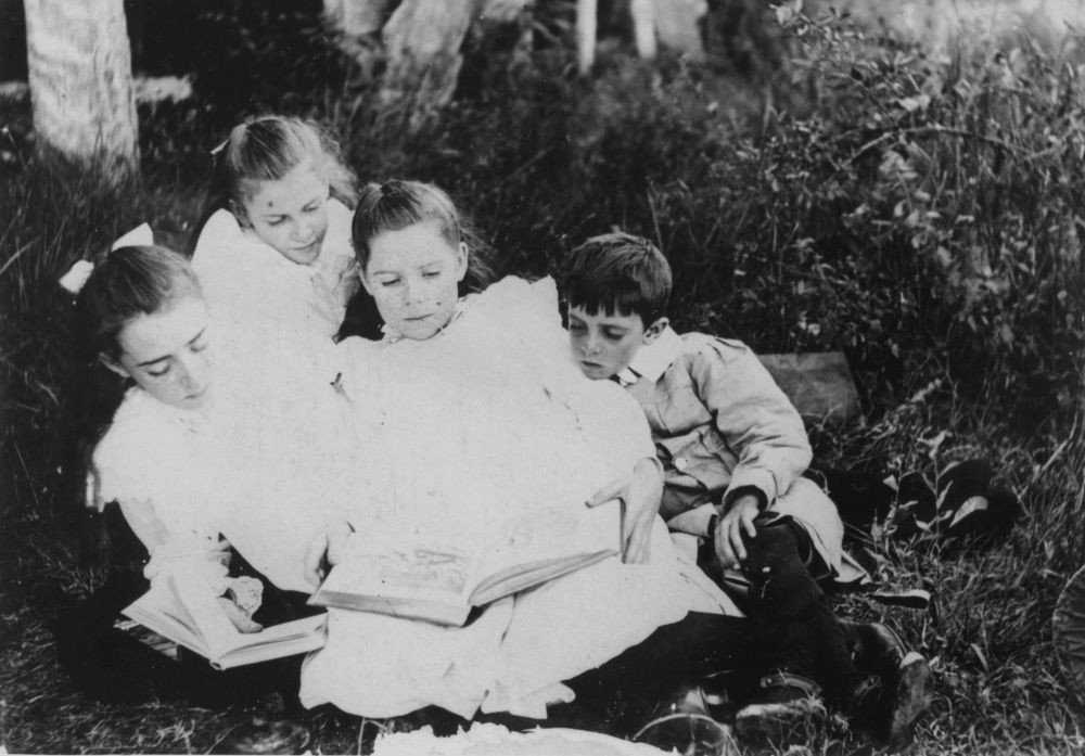 Group of children sitting on the grass reading books, ca.1900-1910 